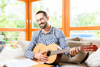 Smiling man playing acoustic guitar indoors, practicing music for brain health
