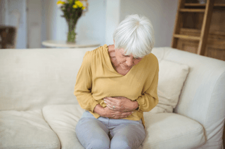 Senior woman sitting on couch holding stomach, appearing to have abdominal discomfort